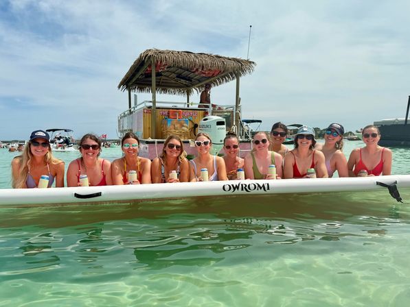 Group of smiling women on a long inflatable float in shallow turquoise water, holding canned drinks with a tiki‑roof party boat in the background on a sunny beach day.