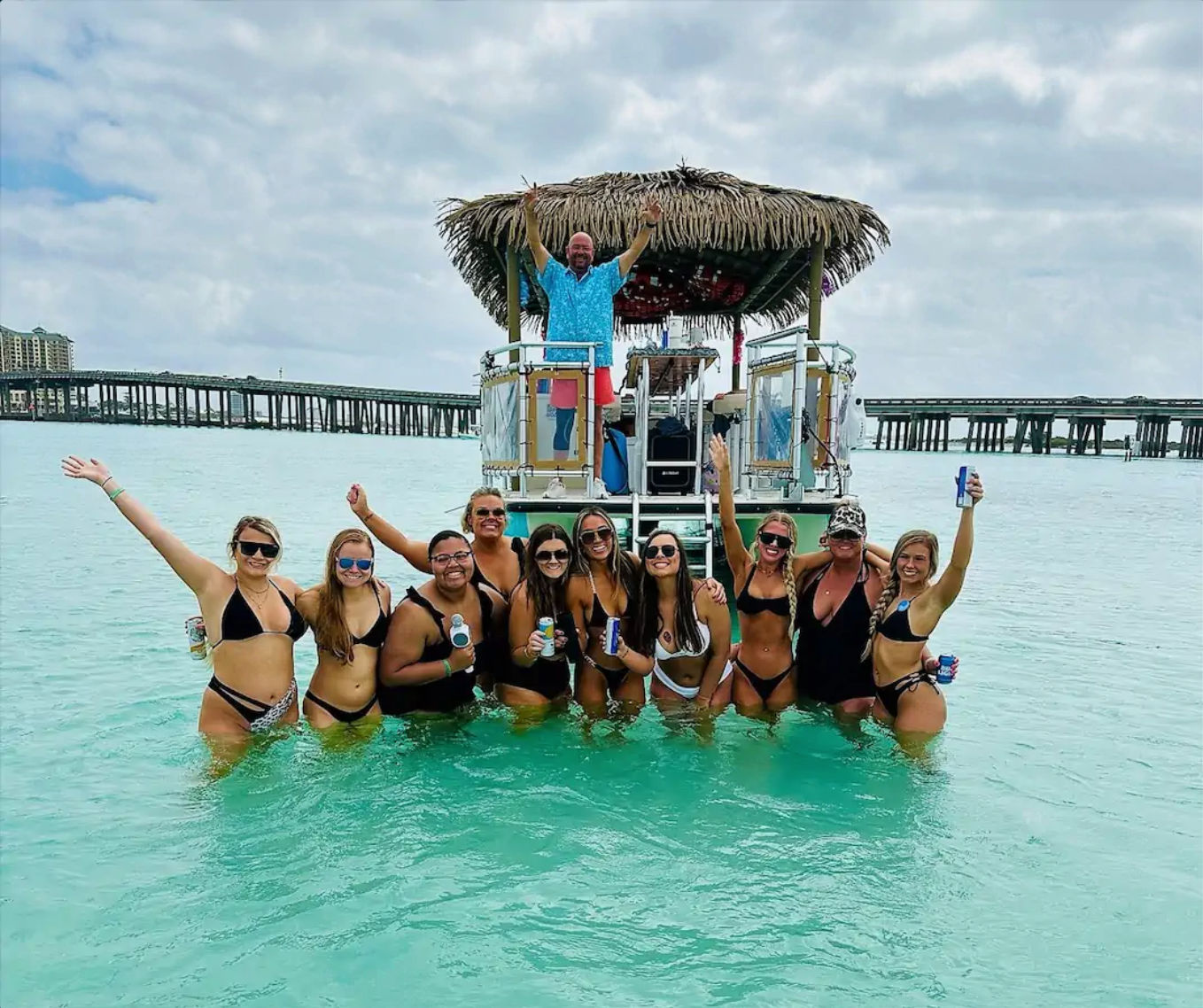 Group of friends in bikinis standing in clear turquoise shallow water, cheering with drinks in front of a thatched-roof party pontoon and a coastal pier under a cloudy sky