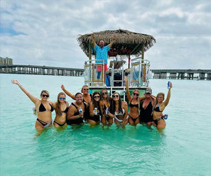 Group of friends in bikinis standing in clear turquoise shallow water, cheering with drinks in front of a thatched-roof party pontoon and a coastal pier under a cloudy sky