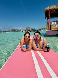 Two smiling women in blue swimsuits and sunglasses lounging on a pink inflatable mat over clear turquoise water, holding canned drinks near a thatched tiki boat and busy beach on a sunny tropical day