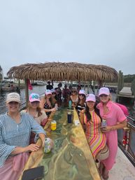 Group of women in colorful summer outfits and pink caps socializing on a tiki-roofed party boat at a marina, seated around a resin-topped bar with drinks, docks and boats in the background.