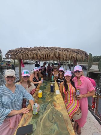 Group of women in colorful summer outfits and pink caps socializing on a tiki-roofed party boat at a marina, seated around a resin-topped bar with drinks, docks and boats in the background.
