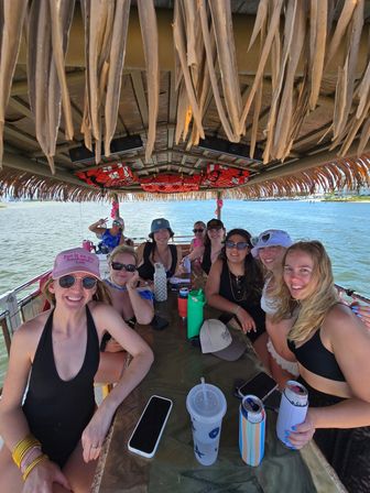 Smiling group of friends on a tiki-style pontoon boat party in a sunny coastal bay, wearing swimsuits and hats with drinks and phones on a central table under a thatched roof
