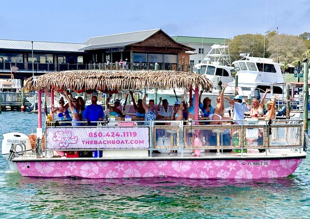 Pink tiki-style party boat with people cheering under a thatched roof, cruising past yachts in a sunny coastal marina.