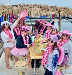 Group of friends on a party boat at a marina, wearing pink cowboy hats, heart-shaped sunglasses, floral leis and tutus around a tiki-style table with drinks