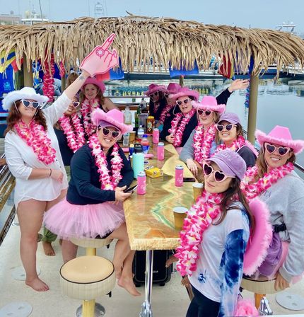 Group of friends on a party boat at a marina, wearing pink cowboy hats, heart-shaped sunglasses, floral leis and tutus around a tiki-style table with drinks