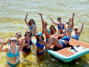 Smiling friends in bikinis and sunglasses waving and holding canned drinks on a floating dock in sunny shallow water