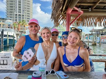 Five friends smiling at a dockside tiki bar in swimwear and sunglasses, tropical thatch roof overhead, boats and a marina with a high-rise in the background; two wear pink SHOWGIRL caps and one holds a cardboard face cutout.