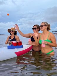 Three women in sunglasses and swimsuits stand waist-deep in calm coastal water around a paddleboard with red cups, tossing an orange ball and holding canned drinks — a playful beach game scene.