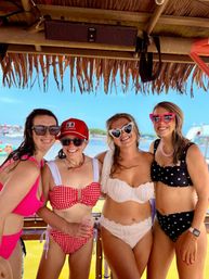 Four women in retro bikinis and heart-shaped sunglasses smiling under a thatched tropical cabana on a sunny beach with blue water in the background.