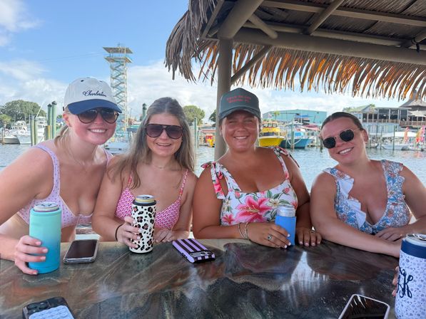 Four friends in swimsuits and sunglasses smiling under a tiki-roof waterfront bar at a marina, holding canned drinks — summer dockside hangout with boats in the background.
