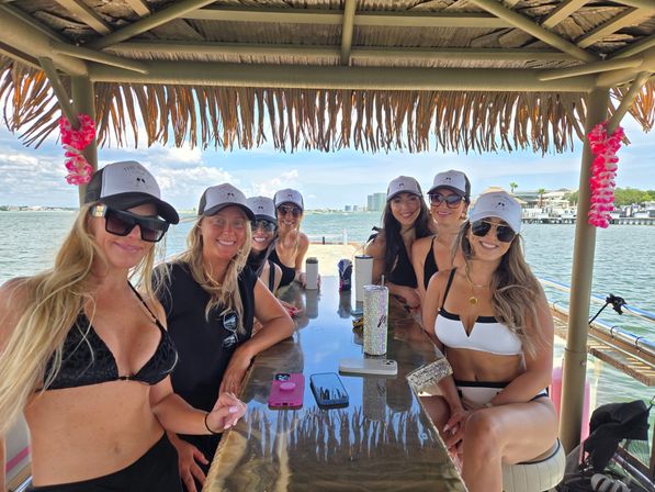 Eight women in swimsuits and matching caps smiling around a shiny bar under a thatched tiki hut on a boat, with sunglasses, drink tumblers and a coastal skyline across calm water.