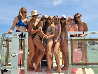 Eight women in colorful swimsuits, sunglasses and a sun hat smiling and posing together on a sunlit boat deck over clear turquoise coastal water
