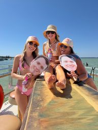Three friends in swimsuits and sun hats smiling on a sunny boat over a lake, wearing sunglasses and holding canned drinks and oversized face cutouts — summer boat party with clear blue sky.