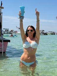 Woman in a light-blue bikini and sunglasses standing waist-deep in clear turquoise water, holding a blue tumbler overhead with boats and a lively sandbar party in the background.