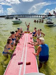 Boat party in a shallow coastal bay — friends in swimsuits gathered around a long pink inflatable table playing a drinking game, anchored boats and a low bridge under dramatic summer clouds.