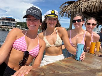 Four smiling young women in summer bikinis at a waterfront tiki bar on a sunny day, one wearing a captain's hat and others holding canned drinks with a marina and docks in the background.