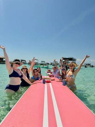 Smiling group of women in swimsuits cheering around a long pink floating dock in shallow turquoise water with anchored boats and a clear blue sky — sunny beach boat party.