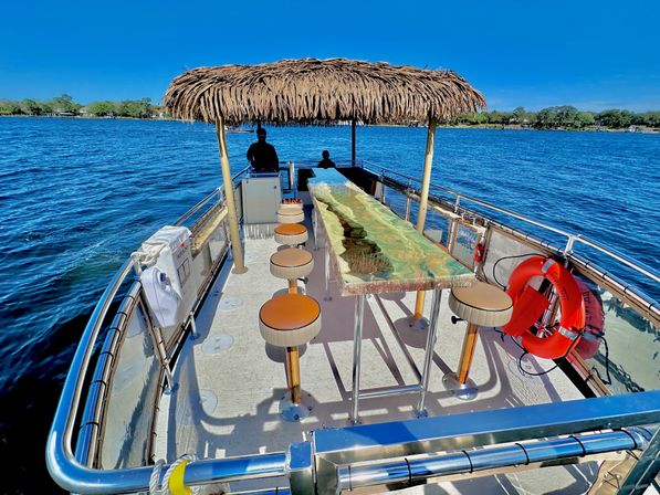 Tiki-roof pontoon bar boat with epoxy resin river-style tabletop, round swivel stools, life rings and two silhouetted passengers on bright blue lake with tree-lined shore