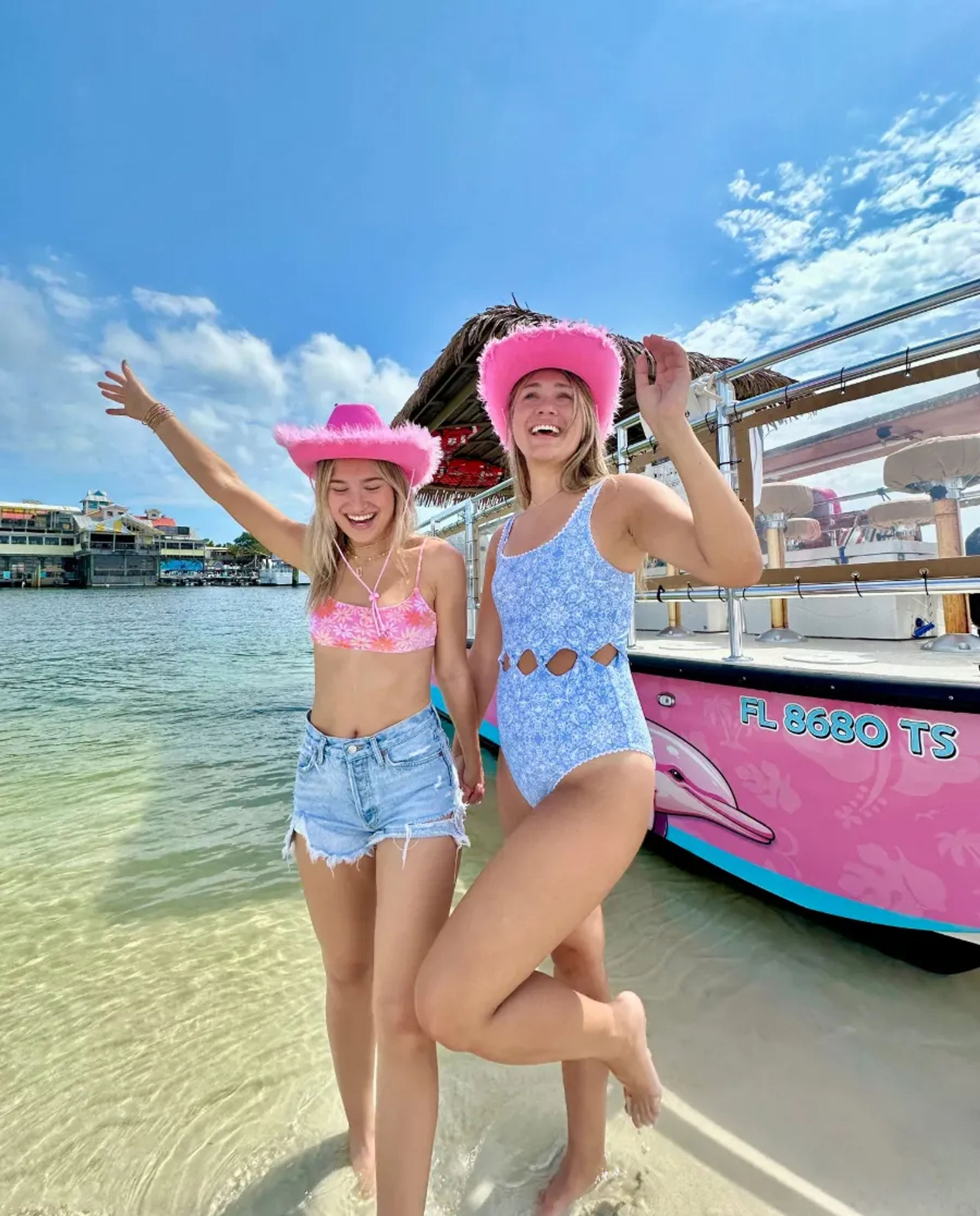 Two women in swimsuits wearing pink fuzzy cowboy hats, laughing in shallow clear water beside a colorful pink boat at a sunny marina with blue skies and waterfront buildings