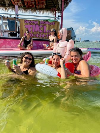 Cheerful group of three people in swimsuits floating on inflatables with canned drinks beside a pink thatched tiki boat and inflatable doll in shallow green bay under a sunny blue sky.