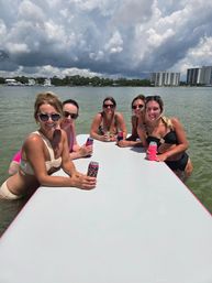Five smiling women in swimsuits gathered around a floating white raft in shallow bay, holding canned drinks with waterfront condos and dramatic cloudy sky in background.