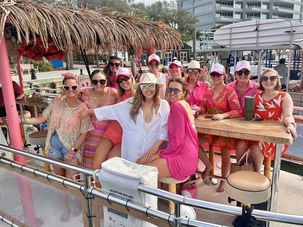 Smiling group of women in pink and summer outfits wearing sunglasses and hats aboard a tiki-roofed pontoon boat at a marina with waterfront condos