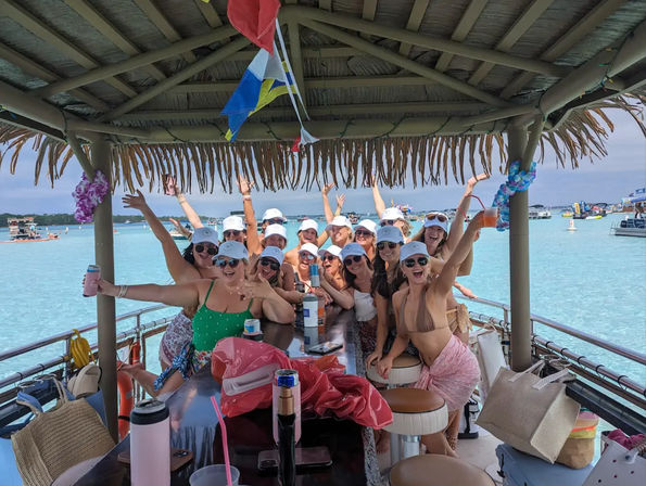 Tiki-roofed pontoon boat party with a cheering group in swimsuits and white caps toasting with drinks over clear turquoise shallow water and other boats in the distance