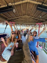 Group of smiling women on a tiki-style boat bar at a marina, raising drinks and waving under a thatched roof with waterfront and boats in the background