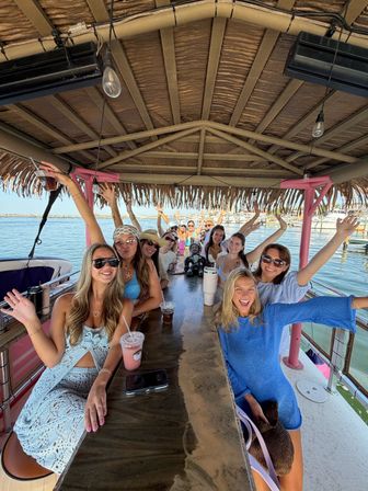 Group of smiling women on a tiki-style boat bar at a marina, raising drinks and waving under a thatched roof with waterfront and boats in the background