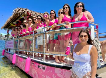 Sunny Florida boat party: a cheerful group of women in pink swimsuits and sunglasses aboard a pink tiki-style party pontoon near the shoreline, one woman in a white swimsuit holding a pink drink can in the foreground.