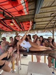 Group of friends in swimsuits laughing at a thatched tiki bar on a tropical beach, woman in a white one-piece posing on the bar under hanging orange life jackets with ocean and distant shoreline in the background.