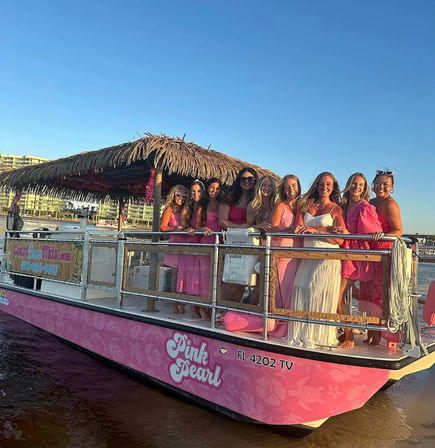 Cheerful group of women in pink and white dresses standing on a pink tiki-style party boat near a Florida shoreline at golden hour, beachfront condos in the background.