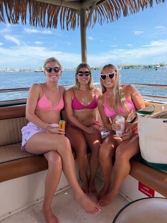 Three friends in pink bikinis smiling on a boat under a thatch canopy, holding canned drinks with a sunny coastal bay and marina in the background — summer boat day.