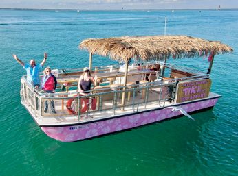 Pink floral tiki pontoon boat with a thatched roof and three people waving on deck, cruising on clear turquoise coastal waters under a sunny sky with a seagull nearby.
