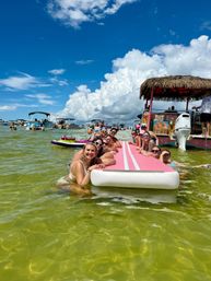 Smiling group of people lounging on a long pink inflatable mat in shallow green coastal water near anchored boats and a tiki-style pontoon under a bright blue sky with puffy white clouds — sunny summer sandbar scene.