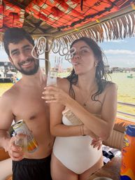 Couple on a sunny beach boat sipping drinks through a long spiral straw under a thatched tiki roof, life jackets overhead and boats and shallow water in the background