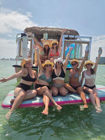 Seven friends in bikinis and straw hats cheering and holding drinks on a floating paddle platform in front of a tiki-roof boat on sunny coastal waters with a distant shoreline and buildings.