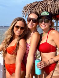 Three smiling women in sunglasses—two in red bikinis and one in a pink polka-dot suit—posing under a thatched tiki umbrella on a sunny tropical beach, one holding a turquoise insulated bottle.