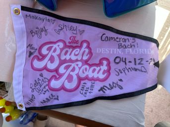 Pink bachelorette party pennant covered in handwritten signatures and a date, printed with “Destin, Florida,” laid on a table next to bottled drinks and a pool bag.