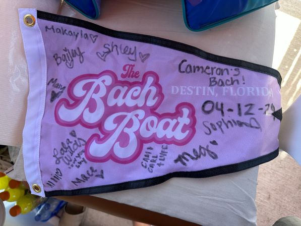 Pink bachelorette party pennant covered in handwritten signatures and a date, printed with “Destin, Florida,” laid on a table next to bottled drinks and a pool bag.