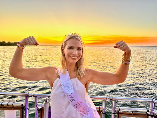 Smiling woman wearing a bridal sash and tiara flexes her arms on a sunset boat cruise over calm water with a golden sky.