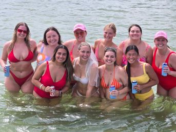 Smiling group of women in colorful bikinis standing waist-deep in calm coastal water, holding canned drinks with a bride in a white bikini and veil at center — fun beach bachelorette party vibe.