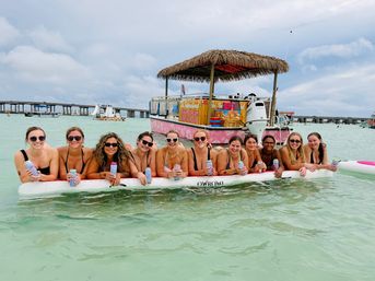 Smiling group of women in swimsuits lounging on an inflatable mat in shallow turquoise water, holding canned drinks with a tiki-roof party boat and low bridge in the background.