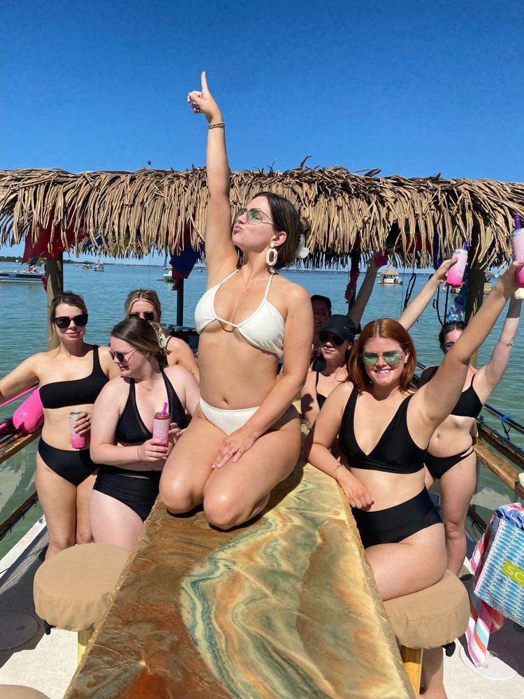 Bikini-clad friends partying on a tiki-roofed boat over turquoise coastal waters, posing and raising drinks under a clear blue sky