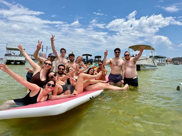 Group of friends in swimsuits cheering and posing on a large paddleboard in shallow coastal water, surrounded by anchored pontoon boats under a sunny blue sky with scattered clouds — lively summer beach day.