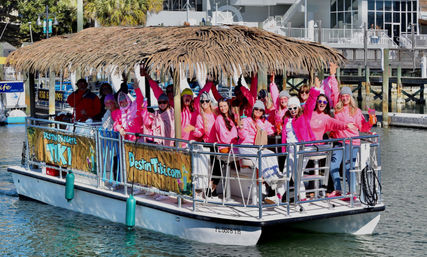 Cheerful group in pink hoodies on a tiki-style pontoon boat cruising a sunny coastal marina