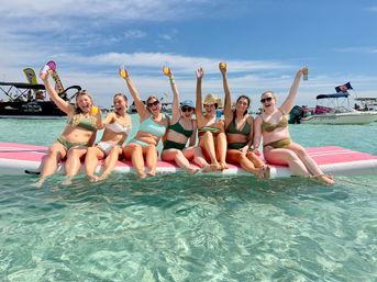 Beach party: eight friends in bikinis sitting on a pink floating mat in clear turquoise shallow water, raising drinks and cheering under a sunny blue sky with boats in the distance.