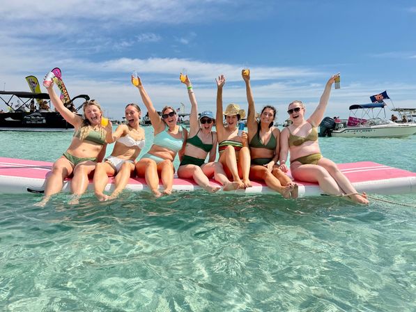 Beach party: eight friends in bikinis sitting on a pink floating mat in clear turquoise shallow water, raising drinks and cheering under a sunny blue sky with boats in the distance.