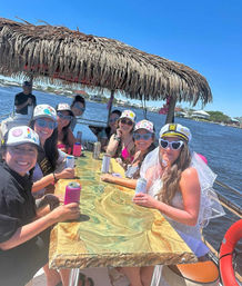 Bachelorette boat party under a tiki-roof on a sunny waterfront—friends in matching caps and heart-shaped sunglasses toast canned drinks around a marbled table; bride-to-be wearing a veil and captain hat.